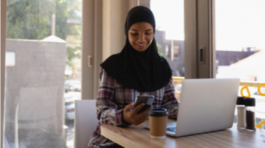 Woman wearing a hijab looking at a phone, sitting with computer at a cafe
